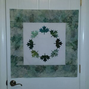 May include: A white door with a green and white fabric wall hanging featuring a wreath of green maple leaves.