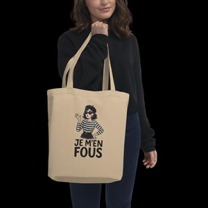Puede incluir: Bolso tote beige con una ilustración de dibujos animados de una mujer con boina, gafas de sol y una camisa a rayas. El texto en francés "JE M'EN FOUS" está impreso debajo. El bolso lo sostiene una persona.