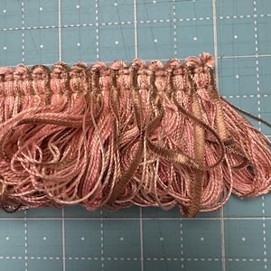 May include: Close-up of a decorative fringe trim. The trim features a combination of pink and gold threads, creating a textured and layered effect. The fringe is likely used for embellishing clothing, home decor, or craft projects.