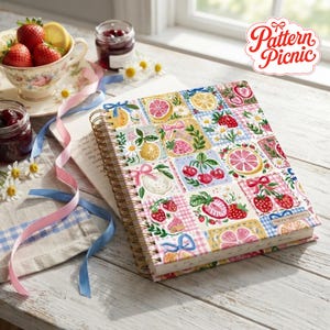 May include: A spiral-bound notebook featuring a colorful grid pattern of strawberries, cherries, citrus slices, daisies, and pink bows. The cover displays the text Pattern Picnic. The notebook is placed on a white wooden surface with ribbons.