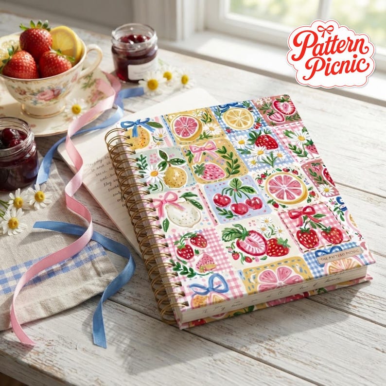 May include: A spiral-bound notebook featuring a colorful grid pattern of strawberries, cherries, citrus slices, daisies, and pink bows. The cover displays the text Pattern Picnic. The notebook is placed on a white wooden surface with ribbons.