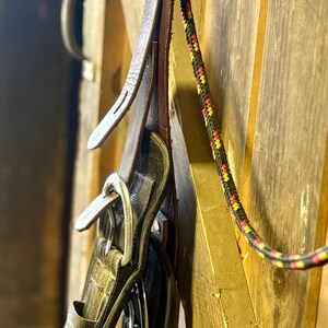May include: Close-up of equestrian equipment hanging on a wooden wall. The image features a leather strap, a black and silver saddle pad, and a multi-colored rope. The items are suspended from a metal ring, suggesting a storage or display setup.