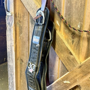 May include: A black horse cinch with silver-colored buckles and the "Z Tech" logo, hanging on a wooden door. The cinch is made of a durable material and features a leather strap and a rope.