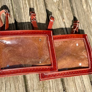 May include: Two red leather saddle bag inserts with clear plastic windows and decorative stitching. Each insert has a leather strap with a buckle for attachment. The inserts are designed to be used on a saddle.