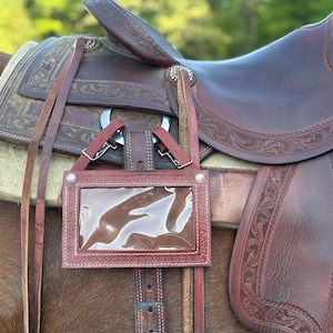 May include: A brown leather saddle with intricate floral tooling and a clear plastic holder. The saddle is attached to a horse and features a silver buckle and various leather straps. The background is blurred with green foliage.
