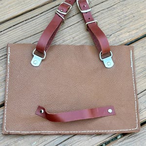 May include: A brown leather portfolio with a textured surface and white stitching. It features two dark brown leather straps with silver buckles and a smaller handle. The portfolio is designed for carrying documents.