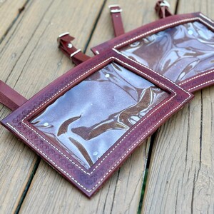 May include: Two burgundy leather name tag holders with clear plastic windows and white stitching. The holders have decorative embossed edges and adjustable leather straps with silver buckles. The items are displayed on a wooden surface.