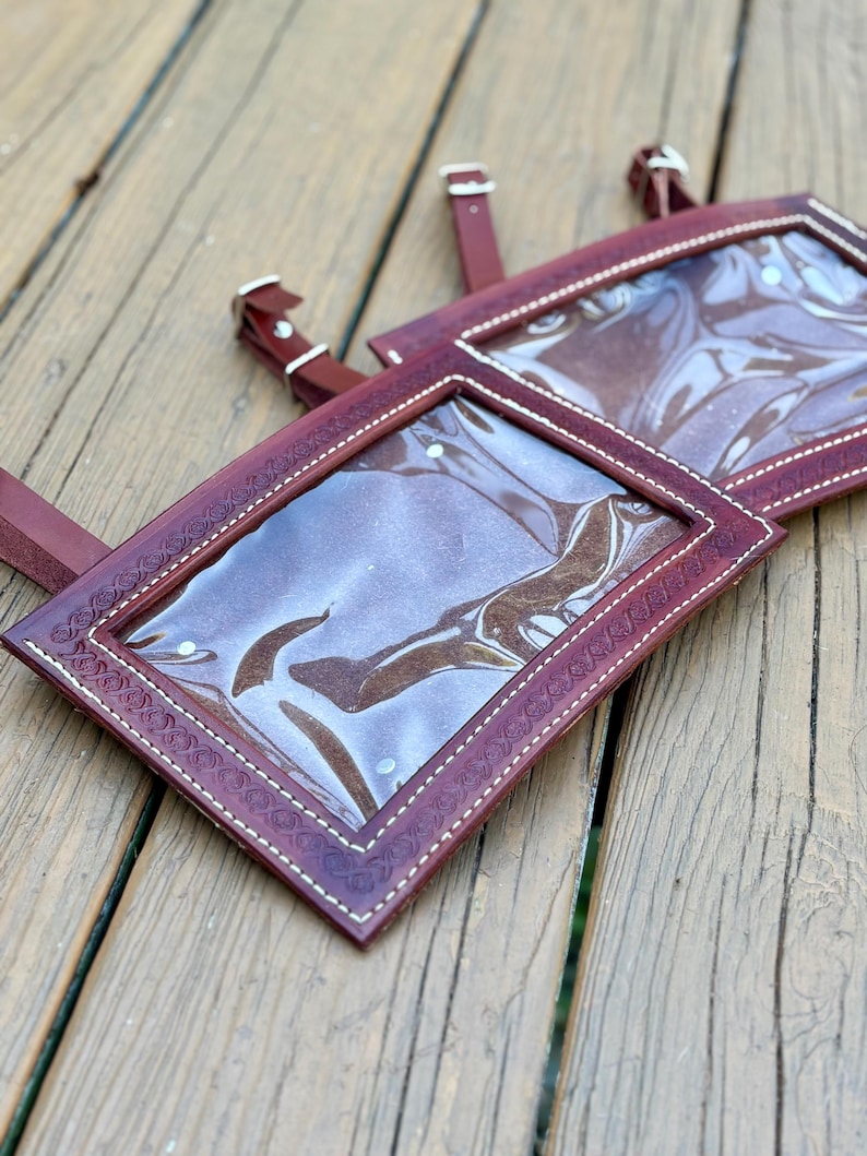 May include: Two burgundy leather name tag holders with clear plastic windows and white stitching. The holders have decorative embossed edges and adjustable leather straps with silver buckles. The items are displayed on a wooden surface.