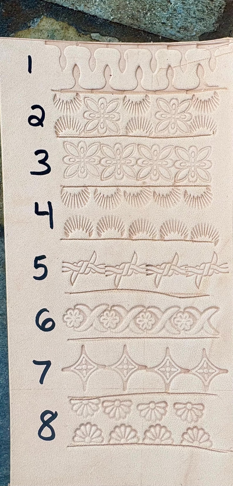 May include: A piece of light tan leather with eight different embossed border designs. The designs include floral patterns, sunbursts, and a barbed wire motif. The numbers 1 through 8 are written in black ink next to each design.