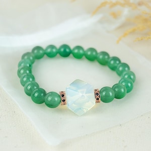 Könnte beinhalten: Ein grünes Perlenarmband mit einem zentralen sechseckigen Opalitstein. Das Armband hat kleine, runde grüne Perlen und ist mit roségoldfarbenen Abstandshaltern mit schwarzen Verzierungen versehen. Das Armband wird auf einer weißen Oberfläche präsentiert.