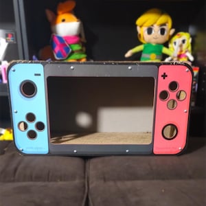 Pode incluir: Um recorte de papelão de um console Nintendo Switch, pintado para parecer um console real. O recorte é azul e vermelho, com uma moldura cinza e botões pretos. O recorte está vazio por dentro, o que sugere que é um adereço ou uma peça de exibição.