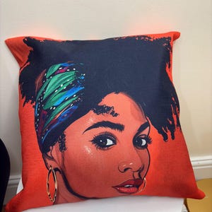 Pode incluir: Uma almofada decorativa com uma ilustração vibrante do rosto de uma mulher. A almofada tem um fundo vermelho com um retrato de uma mulher usando um lenço estampado e brincos de argola dourados. O tom de pele da mulher é marrom avermelhado quente.