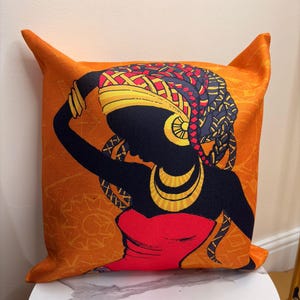 Puede incluir: Cojín decorativo naranja con la silueta de una mujer con un pañuelo en la cabeza y joyas. La silueta de la mujer es negra, con detalles en rojo y dorado. El cojín tiene un patrón geométrico.