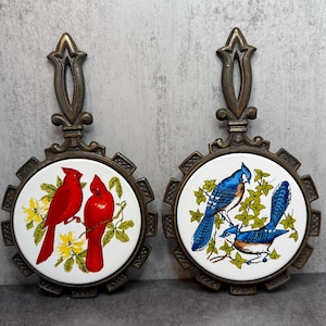 May include: Two vintage trivets with metal handles and decorative ceramic inserts. One trivet features two red cardinals on a white background with yellow flowers. The other has three blue jays with green foliage.
