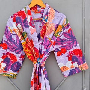 May include: A floral print robe in shades of purple, red, orange, and pink hangs on a wooden hanger. The robe features a tie closure and white trim. The floral pattern includes large leaves and flowers.