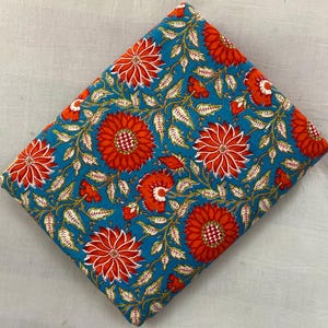 Puede incluir: Tela doblada con un vibrante estampado floral. El diseño presenta grandes flores naranjas con centros blancos y hojas verdes sobre un fondo turquesa. La tela es probablemente de algodón o un material similar.