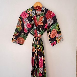 Op de afbeelding: Zwarte badjas met bloemenprint met roze, rode en groene accenten. De badjas heeft een striksluiting en witte bies. Hij hangt aan een houten kleerhanger tegen een witte muur.