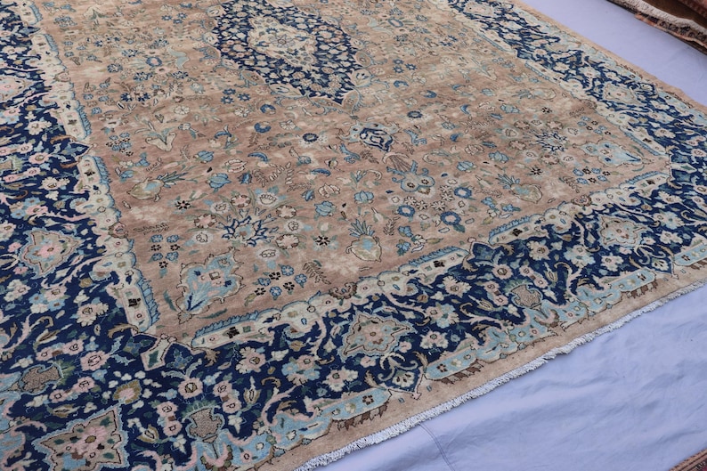 K&ouml;nnte beinhalten: Ein antiker Teppich mit einem beigefarbenen Mittelfeld und einem marineblauen Rand. Der Teppich weist komplizierte Blumenmuster in Blau-, Beige- und Hellgr&uuml;nt&ouml;nen auf. Das Design umfasst ein zentrales Medaillon und Eckakzente.