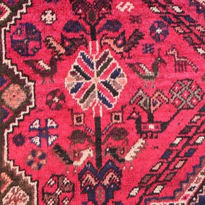 May include: Close-up of a vibrant red oriental rug with intricate patterns. The rug features a central floral design, stylized birds, and geometric borders in shades of blue, brown, and white. The rug's texture is visible.
