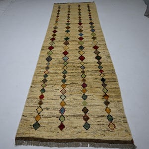 Puede incluir: Una alfombra pasillera larga y rectangular con una base beige texturizada. La alfombra presenta cuatro columnas verticales de pequeños rombos multicolores en rojo, azul, verde, amarillo y marrón. Los extremos tienen flecos marrones.