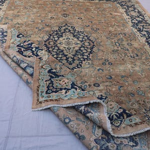 K&ouml;nnte beinhalten: Ein antiker Teppich mit einem zentralen Medaillon-Design in Marineblau und Hellblau auf beigem Hintergrund. Der Teppich hat ein abgenutztes, Vintage-Aussehen mit aufwendigen floralen Mustern und einem Fransenrand. Der Teppich ist teilweise gefaltet und zeigt die Unterseite.