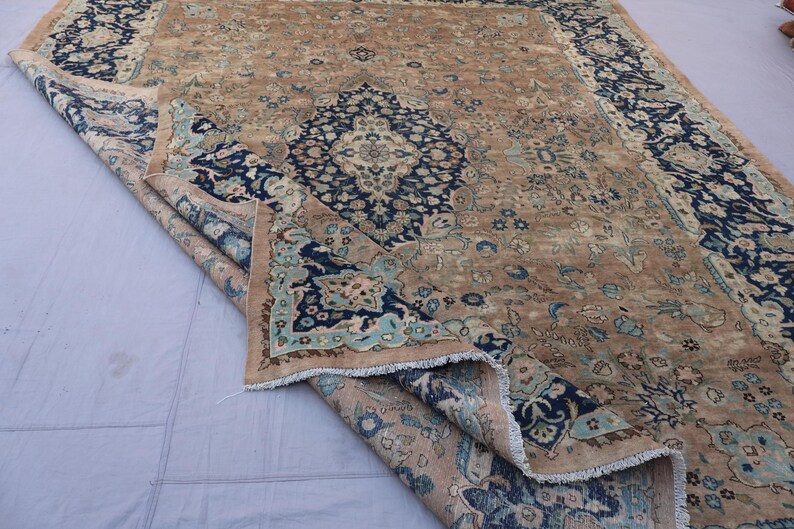 K&ouml;nnte beinhalten: Ein antiker Teppich mit einem zentralen Medaillon-Design in Marineblau und Hellblau auf beigem Hintergrund. Der Teppich hat ein abgenutztes, Vintage-Aussehen mit aufwendigen floralen Mustern und einem Fransenrand. Der Teppich ist teilweise gefaltet und zeigt die Unterseite.