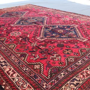 May include: A large, rectangular red rug with intricate geometric patterns and a dark blue central design. The rug features a detailed border with cream, brown, and black accents, and a fringed edge. The rug is likely handmade.