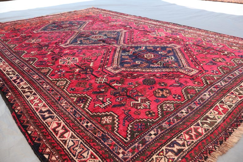 May include: A large, rectangular red rug with intricate geometric patterns and a dark blue central design. The rug features a detailed border with cream, brown, and black accents, and a fringed edge. The rug is likely handmade.
