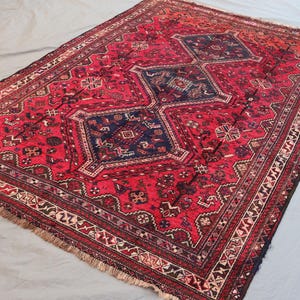 May include: A rectangular, hand-woven rug with a red field and intricate geometric patterns. The rug features a central design of three dark blue diamond shapes, surrounded by detailed borders in various colors. The rug has a fringed edge.