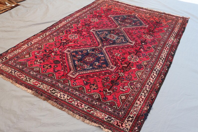 May include: A rectangular, hand-woven rug with a red field and intricate geometric patterns. The rug features a central design of three dark blue diamond shapes, surrounded by detailed borders in various colors. The rug has a fringed edge.