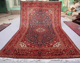Puede incluir: Una gran alfombra persa rectangular con un diseño de medallón central. La alfombra presenta un borde rojo intenso y un campo azul marino lleno de intrincados patrones florales. Flecos blancos bordean la alfombra.
