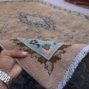 K&ouml;nnte beinhalten: Ein Abschnitt eines beige-blauen gemusterten Teppichs wird hochgehalten und zeigt ein detailliertes Eckdesign. Der Teppich hat einen Used-Look mit einer hellblauen und braunen Farbpalette. Der Fransenrand ist wei&szlig;.