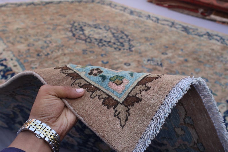 K&ouml;nnte beinhalten: Ein Abschnitt eines beige-blauen gemusterten Teppichs wird hochgehalten und zeigt ein detailliertes Eckdesign. Der Teppich hat einen Used-Look mit einer hellblauen und braunen Farbpalette. Der Fransenrand ist wei&szlig;.