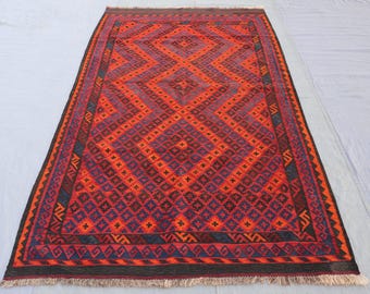 May include: A rectangular rug with a geometric diamond pattern in shades of red, orange, and purple. The rug has a dark border and fringe along the edges. The design features repeating diamond shapes.