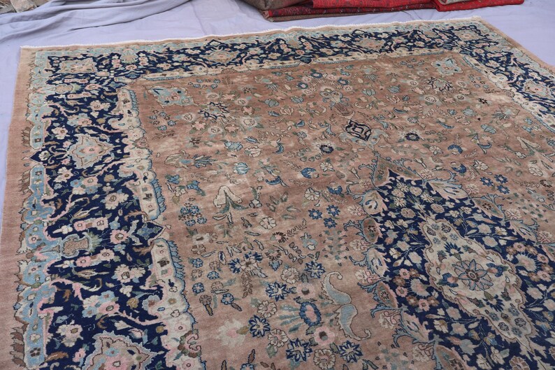 K&ouml;nnte beinhalten: Ein antiker persischer Teppich mit einem zentralen Feld in Beige und aufwendigen floralen Mustern in Blau-, Rosa- und Cremet&ouml;nen. Der Rand ist ein tiefes Marineblau mit floralen Motiven. Das Design des Teppichs ist symmetrisch und detailliert.