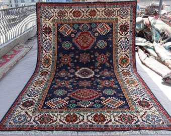 May include: A large, rectangular rug with a navy blue field and intricate geometric patterns in red, orange, green, and cream. The rug features a detailed border and fringe along the edges, showcasing a traditional design.