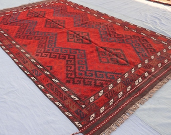 May include: A rectangular red rug with a geometric pattern in dark blue and brown. The rug has a detailed border with white and brown accents and fringe on the edges. The rug is likely made of wool or a similar material.