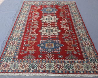 May include: A rectangular red rug with an intricate geometric pattern. The rug features a central design with blue and white diamond shapes, surrounded by a border of repeating patterns in various colors. The rug has a fringe along the edges.