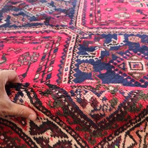 May include: A close-up of a hand holding a section of a vibrant, patterned rug. The rug features a rich color palette of deep red, navy blue, and touches of brown and beige. The intricate design includes geometric shapes and floral motifs, creating a traditional aesthetic.