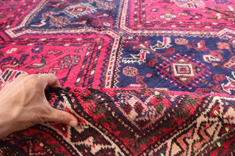 May include: A close-up of a hand holding a section of a vibrant, patterned rug. The rug features a rich color palette of deep red, navy blue, and touches of brown and beige. The intricate design includes geometric shapes and floral motifs, creating a traditional aesthetic.