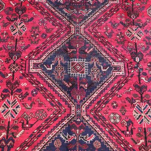 May include: A detailed, close-up view of a red and navy blue oriental rug. The rug features a central diamond pattern with intricate geometric and floral designs. The border has a repeating pattern in shades of red, navy, and cream.