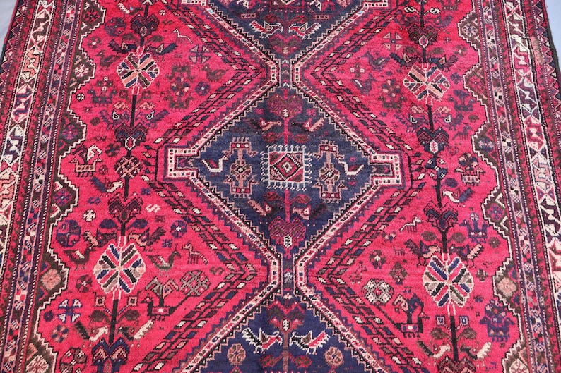 May include: A detailed, close-up view of a red and navy blue oriental rug. The rug features a central diamond pattern with intricate geometric and floral designs. The border has a repeating pattern in shades of red, navy, and cream.