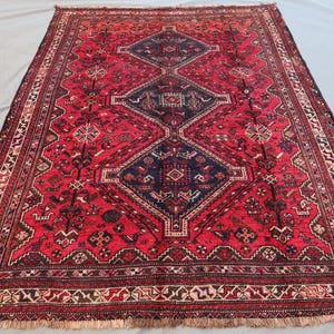 May include: A rectangular, red oriental rug with a central diamond pattern in navy blue. The rug features intricate geometric designs and a detailed border. Fringes are visible along the edges. The rug is a traditional home decor item.