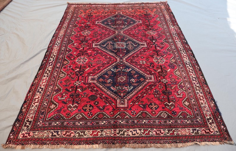 May include: A rectangular, red oriental rug with a central diamond pattern in navy blue. The rug features intricate geometric designs and a detailed border. Fringes are visible along the edges. The rug is a traditional home decor item.