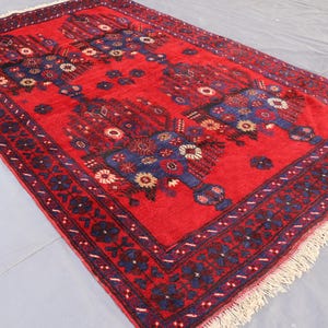 May include: A rectangular red rug with a detailed floral pattern in blue, white, and yellow. The rug has a dark blue border with floral designs and a fringed edge. The rug is made of a soft, woven material.