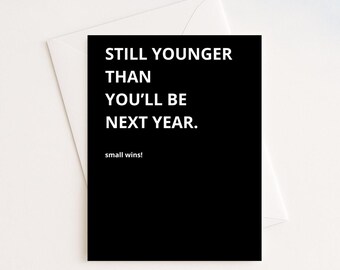 May include: A black greeting card with white text that reads "STILL YOUNGER THAN YOU'LL BE NEXT YEAR." The phrase "small wins!" is below the main text. The card is inside a white envelope.