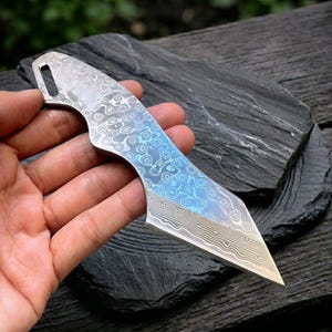 May include: A Damascus steel knife with a distinctive pattern and a blue and silver colour scheme. The blade has a pointed tip and a curved handle with a lanyard hole. The knife is held in a hand.