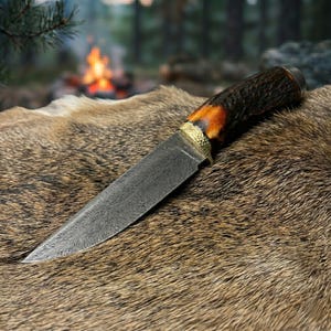 May include: A hunting knife with a patterned steel blade and a handle made of dark brown and orange material. The knife has a gold-colored bolster and rests on a brown fur surface. The background shows a blurred forest and a campfire.