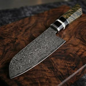 May include: A Damascus steel chef's knife with a patterned blade and a handle featuring black, silver, and green accents. The knife rests on a dark wooden cutting board, showcasing its intricate design and craftsmanship.