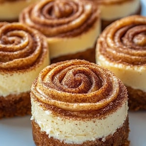 Pode incluir: Close-up de vários mini cheesecakes de rolinhos de canela. Cada sobremesa tem uma base de biscoito, um recheio cremoso de cheesecake e uma cobertura de redemoinho de canela. As sobremesas são dispostas em um prato branco.